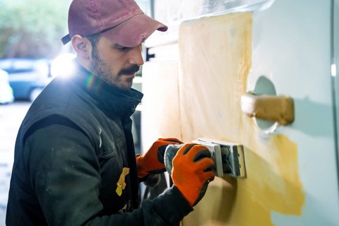 Skilled auto body worker wearing work gloves and a cap carefully sanding a car door with filler