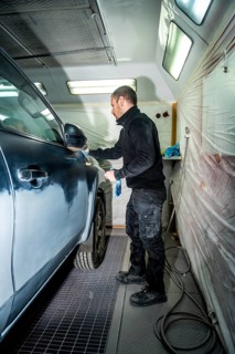 Auto body technician cleaning and preparing a dark colored car for painting inside a professional