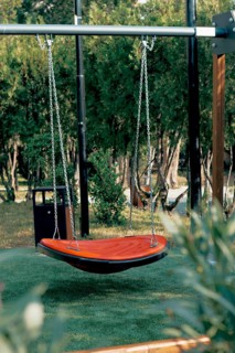 A bright orange hanging swing is set in a serene garden surrounded by trees and grass. The peaceful
