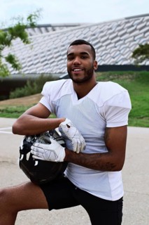 A confident American football player in uniform poses outdoors, holding his helmet. The setting