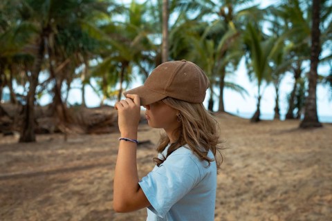 A young woman wearing a casual brown cap and blue shirt stands on a sandy beach, surrounded by lush