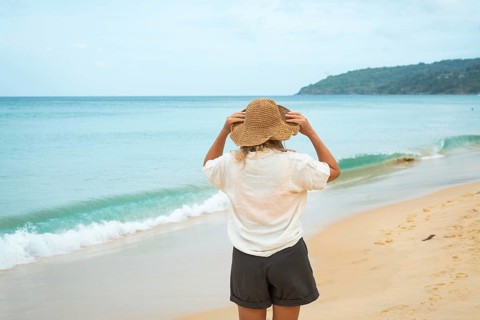 A woman, wearing a sun hat, stands on a sandy beach, gazing at the calm ocean. The scene captures a