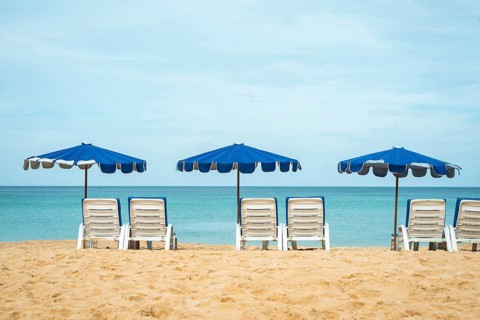A serene beach scene shows neatly aligned lounge chairs beneath blue umbrellas facing a tranquil