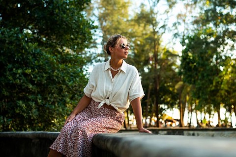 A stylish woman in sunglasses and summer attire sits on a stone ledge surrounded by lush greenery