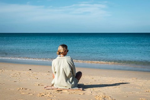 A woman sits calmly on a sandy beach, gazing at the serene ocean under a clear sky in Thailand. Her