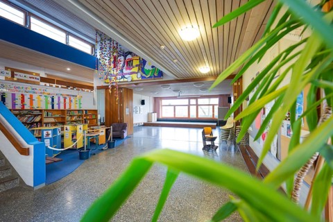 A bright school corridor with works of art, bookshelves and numerous plants in front of large