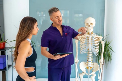 Physiotherapist in purple scrubs pointing at a human skeleton model to a female patient, explaining
