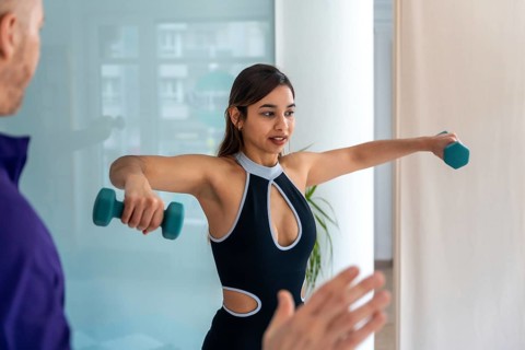 Young woman doing lateral dumbbell raises with a physiotherapist in clinic, focused on shoulder
