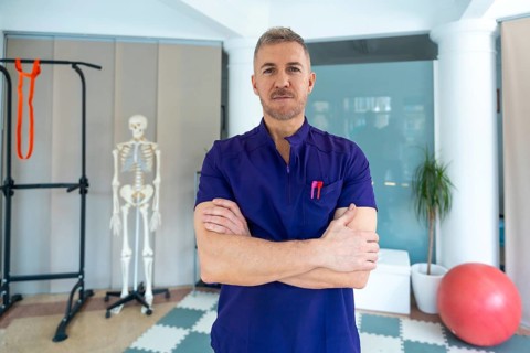 Confident male physiotherapist or osteopath standing with arms crossed in clinic, providing