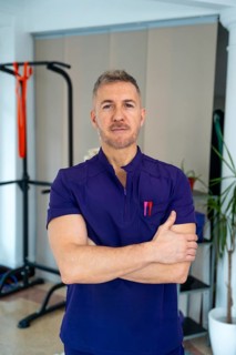 Male physiotherapist in purple uniform stands with arms crossed in a clinic, representing concepts