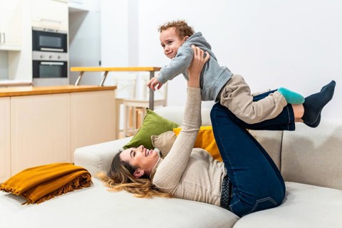 Mother lying on a sofa at home, joyfully playing with her young son, lifting the toddler up in the