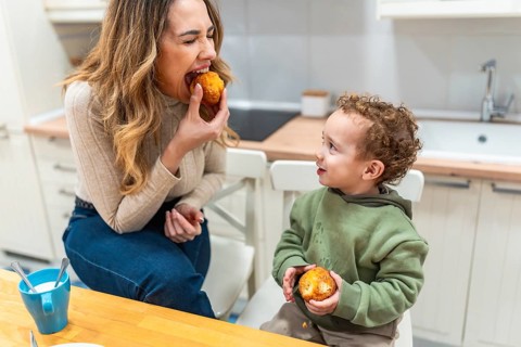 Mother and young son sit at a bright kitchen counter sharing muffins and morning coffee, enjoying a