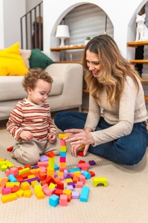 Mother and young son are sitting on the floor at home, building a tower with vibrant wooden blocks,