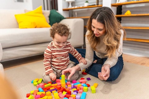 Loving mother and her smiling toddler son are sitting on the floor at home, engaging in creative