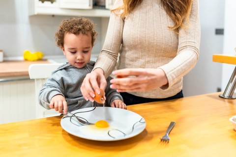 Mother teaching young child to crack an egg and prepare ingredients at the kitchen counter, bonding