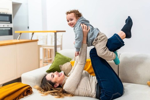 Mother lying on couch lifts smiling toddler in playful airplane pose, sharing a cozy, candid