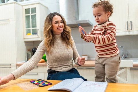 Mother and young child bonding over a coloring book and watercolor palette on a kitchen table,