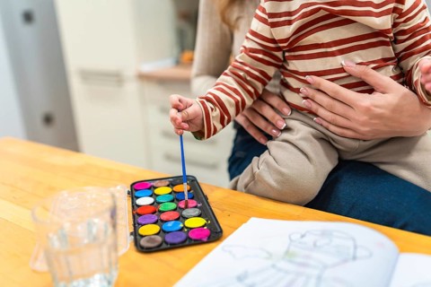 Young child held by mother, dipping a brush into a vibrant watercolor palette while coloring in an