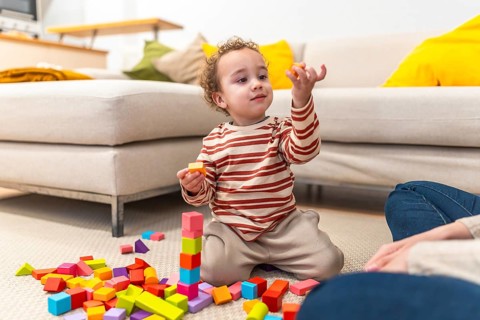 Toddler boy concentrating as he builds a tall wooden block tower on the living room carpet while an