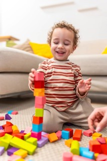 Happy toddler sits on carpet building a tall colorful wooden block tower, smiling and engaged in