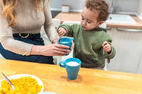 Mother guiding her toddler son stirring milk in a cup during breakfast, creating a moment around
