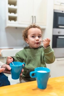 Curly haired toddler sitting at a kitchen table, independently eating yogurt from a spoon, leaving
