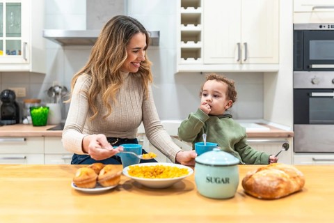 Young smiling mother feeding cereal to her adorable toddler son while sitting at the kitchen table,