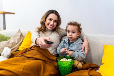 Mother and child relaxing on a cozy sofa covered by a blanket, enjoying popcorn and bonding