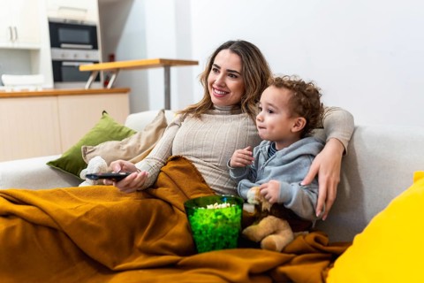 Mother and young child cozy on the sofa sharing popcorn and a remote, smiling and watching tv