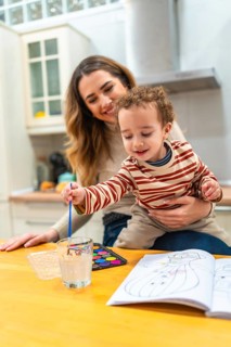 Happy mother holding her young son who is attentively painting watercolors in a coloring book,