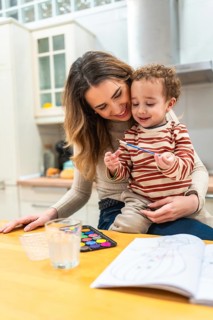 Mother holding her toddler son while they are painting with watercolors at a wooden table in a