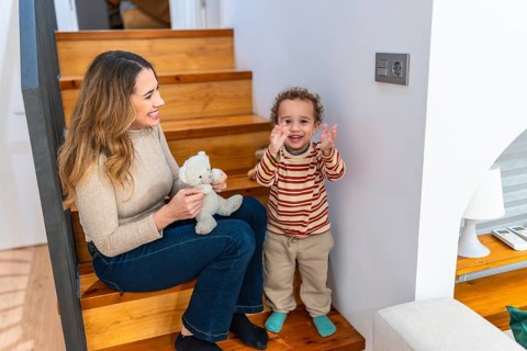 Mother and young son sit on wooden stairs at home, playing with a teddy bear and sharing a warm,