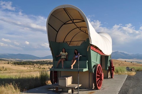 Two people sitting in a huge covered wagon, Conestoga wagon, with mountains in the background,