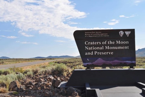 Craters of the Moon National Monument sign next to a road with wide landscape and blue sky, Craters