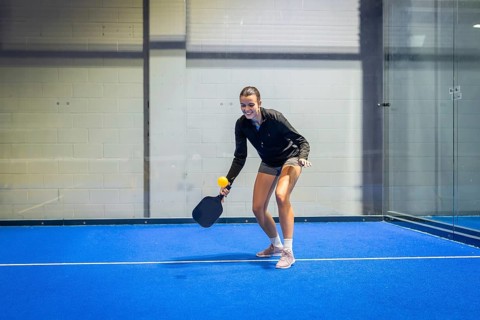Woman athlete smiling, holding a paddle and yellow ball on a vibrant blue pickleball court,