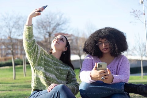 Two friends in the park, one takes a selfie while the other looks at her phone. They are enjoying a