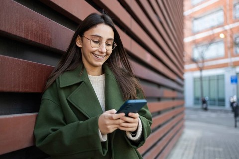 A woman wearing a stylish green coat leans against a wooden wall while engaging with her smartphone
