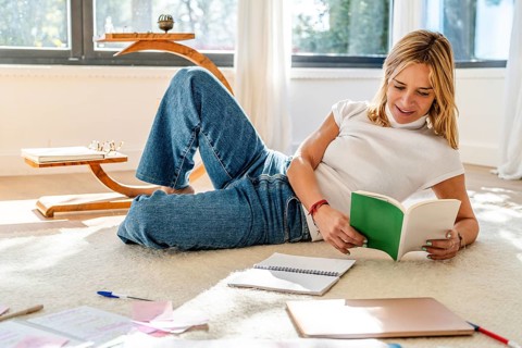 A woman enjoys reading a book while relaxing on a carpeted floor, surrounded by notebooks and a
