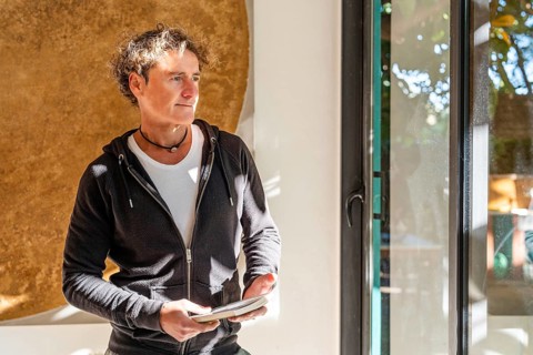 A relaxed man stands indoors holding a book, bathed in natural sunlight near a window Wearing a