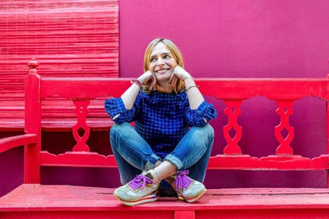 A cheerful young woman in a blue plaid shirt is seated on a bright red bench, smiling and looking