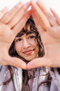 A young Latin woman with curly hair smiles through a hand frame, enjoying a sunny day Her casual