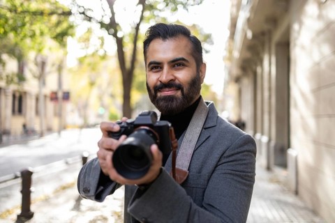Indian male photographer holding a camera, smiles while looking away, standing on a sidewalk lined