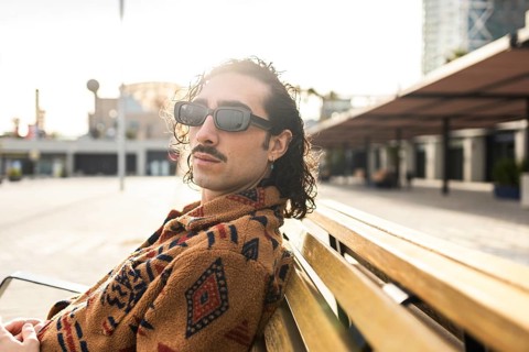 Fashionable man with curly hair and sunglasses lounges on a wooden bench in an urban setting The