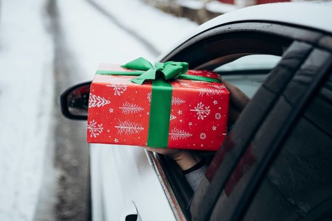 A hand extends a beautifully wrapped gift with red and green holiday paper from a car window The