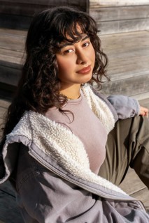 A young Latin woman with curly hair sits on wooden steps, enjoying a sunny day She wears a cozy