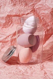 Soft pastel cosmetic sponges in a transparent jar set against a crumpled pink paper background This
