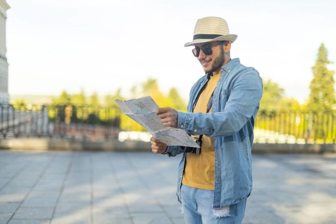A Latino transgender man, wearing casual denim and a straw hat, navigates a city with a map in hand