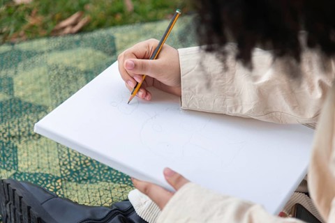 A young black woman sketches on a blank canvas using a pencil, surrounded by grass and natural