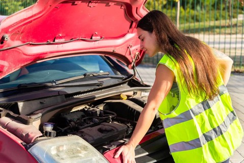 A woman in a reflective safety vest inspects a car engine by the roadside, with the hood open,
