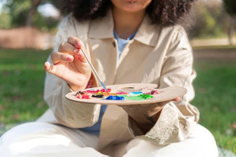 A young black artist holds a paintbrush and a palette filled with vibrant colors, engaging in plein